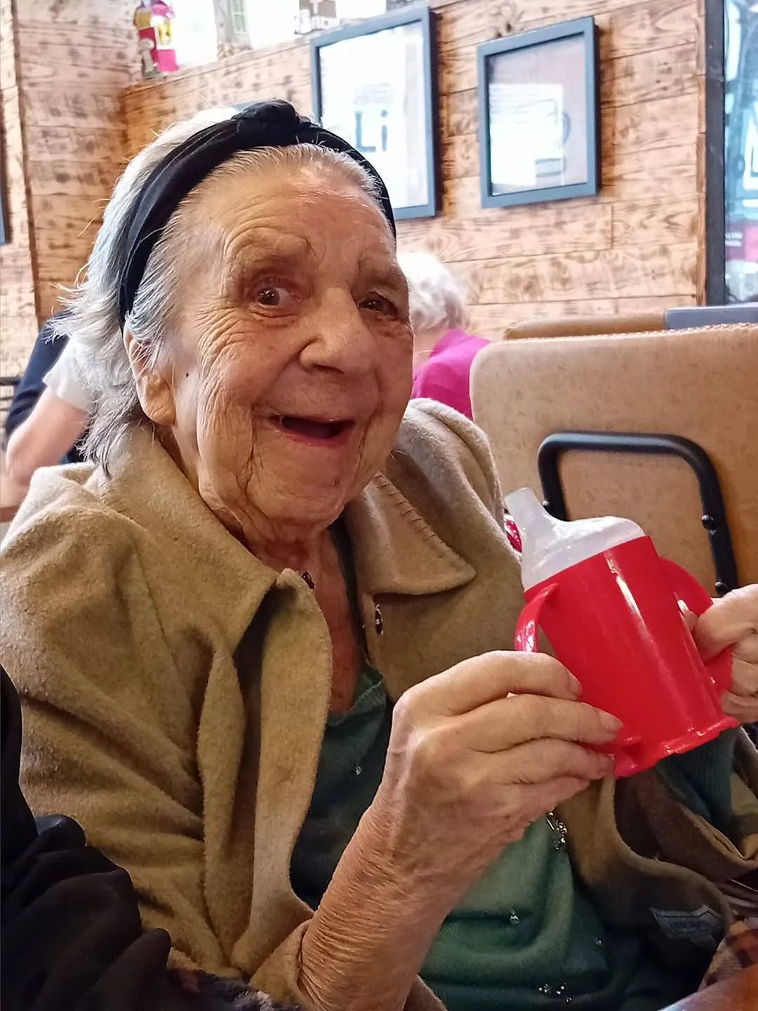 Smiling elderly woman holding a red handled cup, enjoying a warm moment in a cosy Astar Homecare setting.