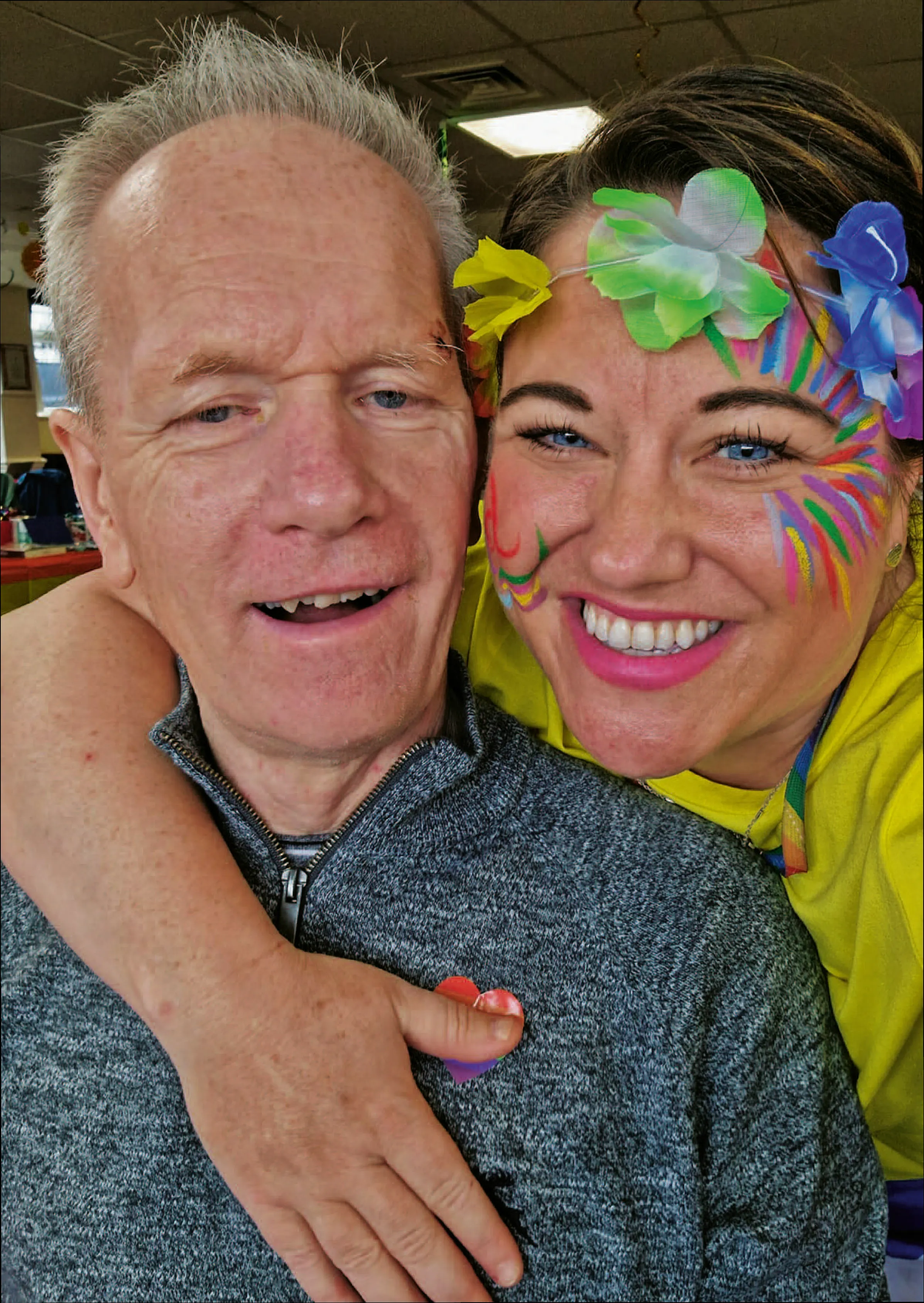 Carer with colourful face paint hugging a smiling male client during a cheerful Astar Homecare event.