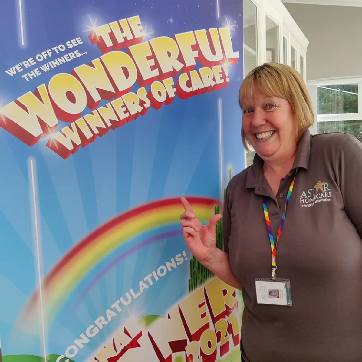Astar Homecare staff member posing beside a ‘Winners of Care’ celebration banner with a rainbow design.