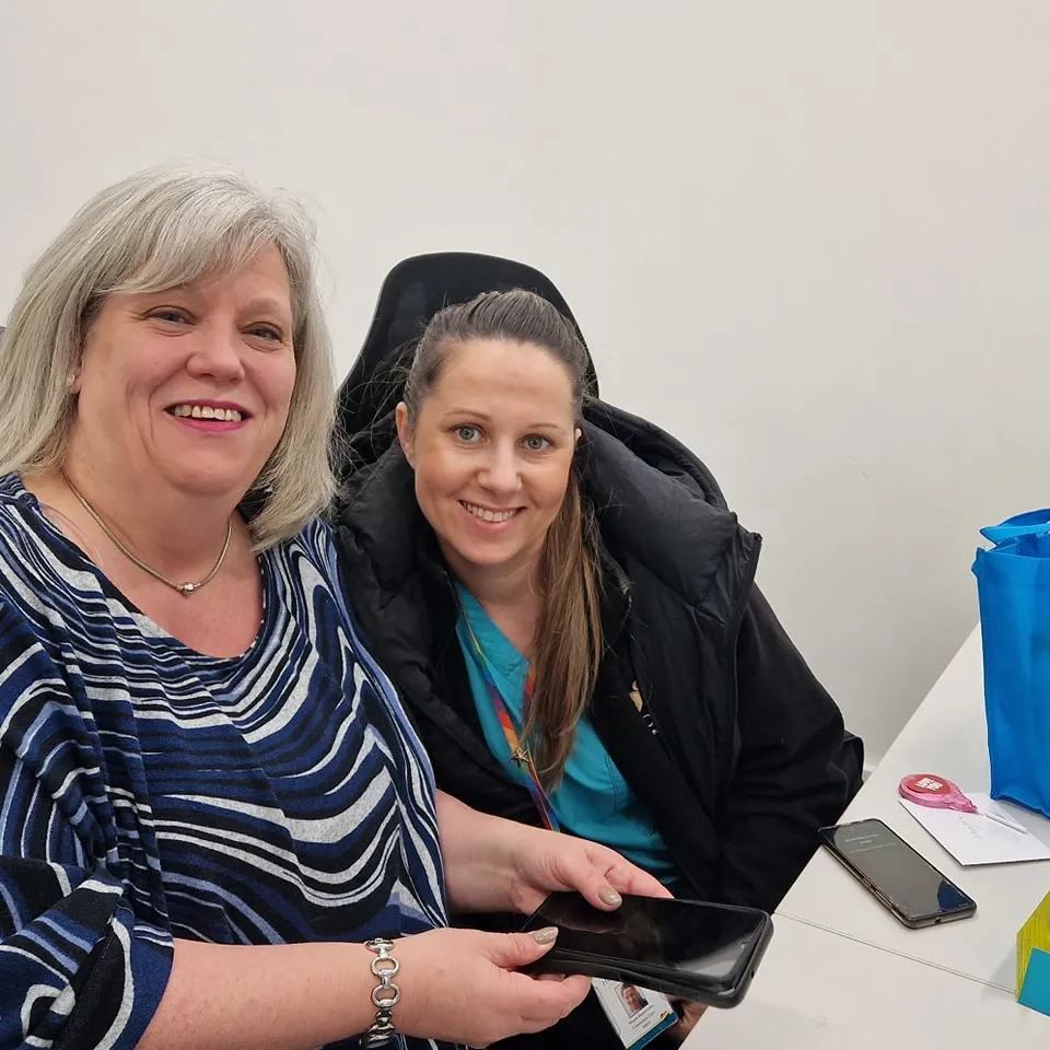 Two women, including an Astar Homecare carer in teal scrubs, smiling at a table with a smartphone.