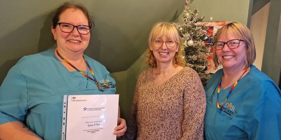 Two Astar Homecare carers and a colleague posing in front of a decorated Christmas tree with a certificate.