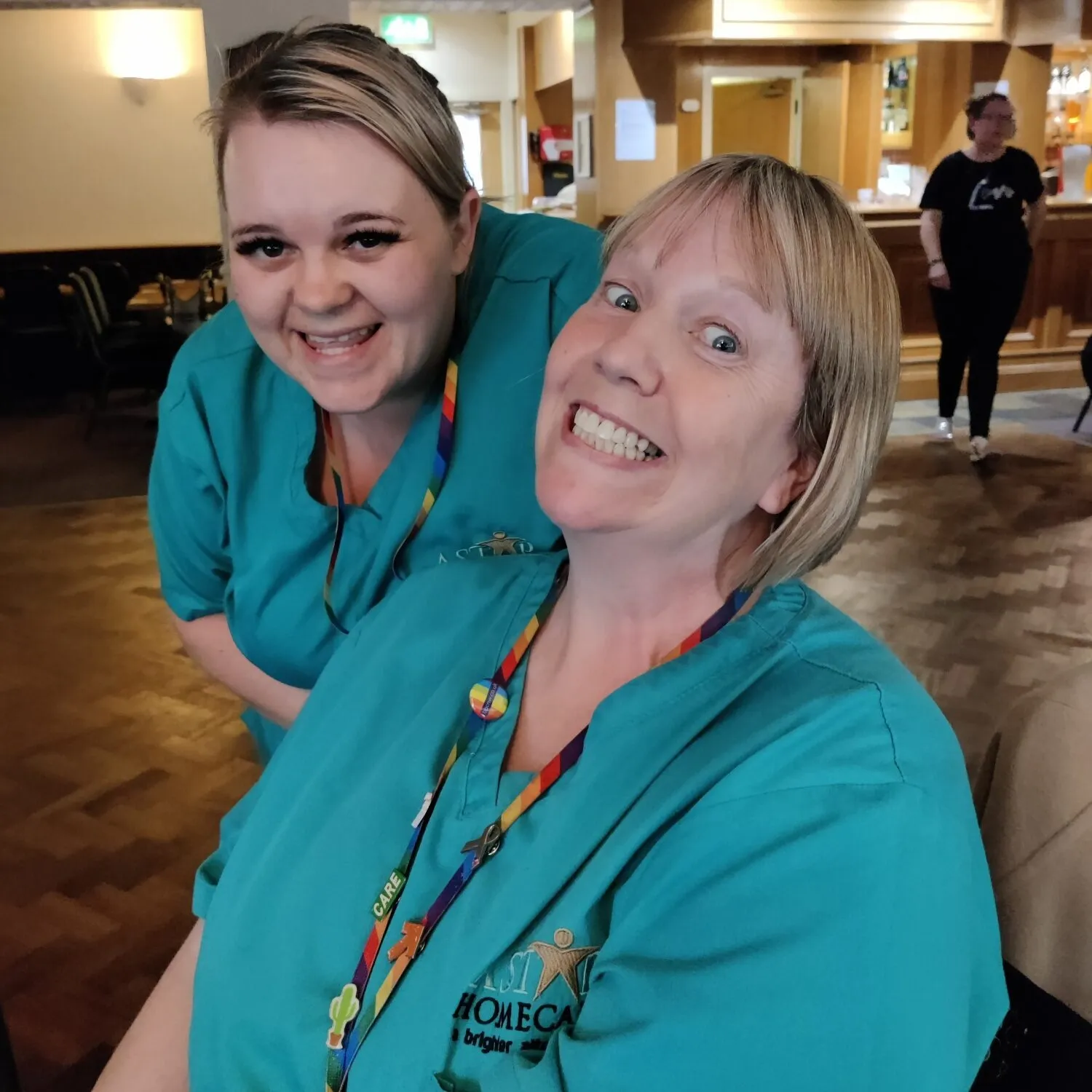 Two smiling Astar Homecare carers posing for a cheerful indoor photo in teal uniforms with rainbow lanyards.