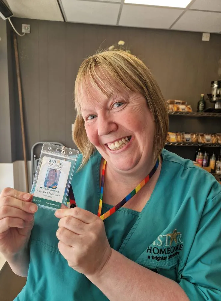 Astar Homecare carer proudly holding her staff ID badge with a big smile in a coffee shop.