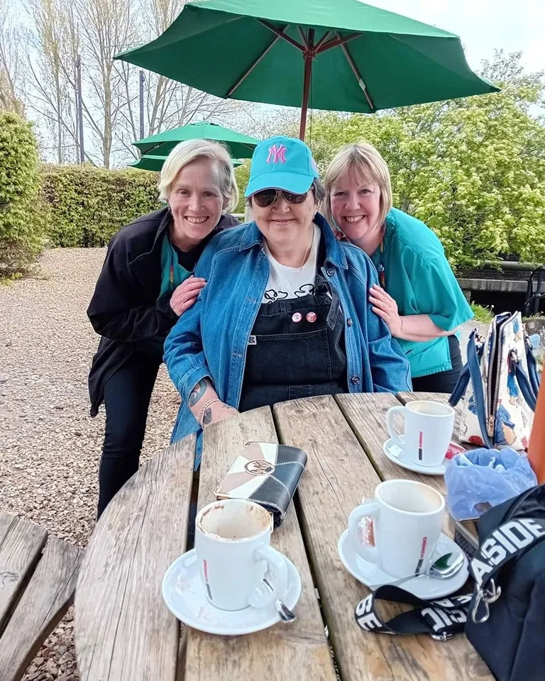 Two Astar Homecare carers sharing a joyful outdoor moment with a client at a café table with coffee.