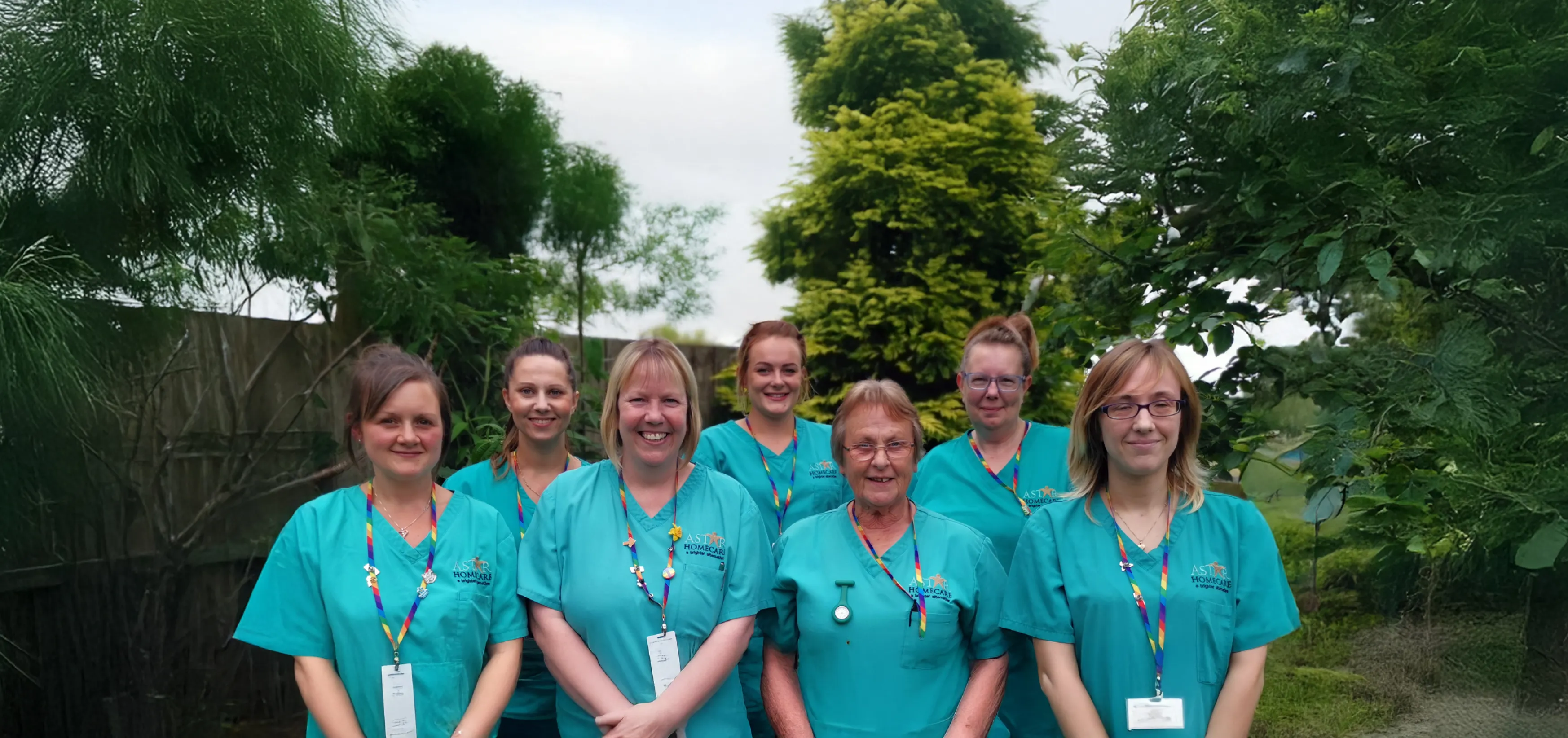 Group photo of eight smiling Astar Homecare team members in turquoise uniforms, standing together outdoors in a garden setting.